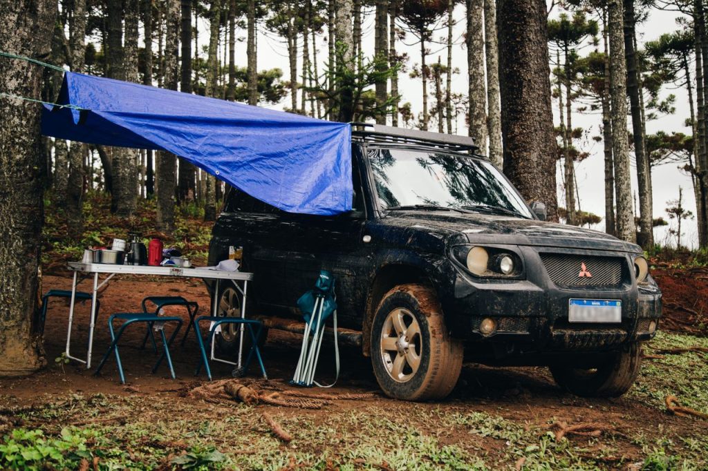 SUV parked in a forest with camping setup, including a tarp and folding chairs, offering an outdoor adventure feel.